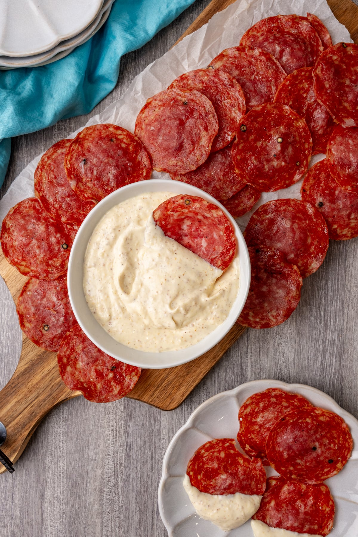 Crispy salami chips arranged around a white bowl of creamy dip on a wooden board with parchment paper and a blue napkin
