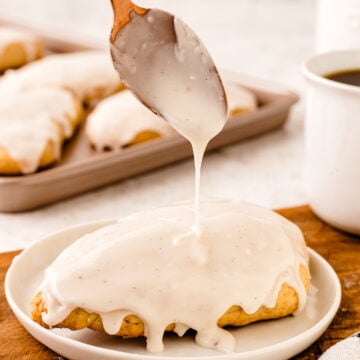 Wooden spoon drizzling vanilla glaze over a warm vanilla scone on a white plate, with more scones blurred in background