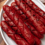 Plate of six BBQ hotdogs with charred skin and grill marks on white plate, wooden surface background with beer bottle visible