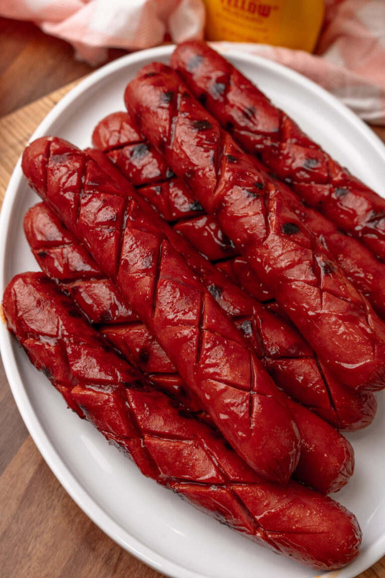 Plate of six BBQ hotdogs with charred skin and grill marks on white plate, wooden surface background with beer bottle visible
