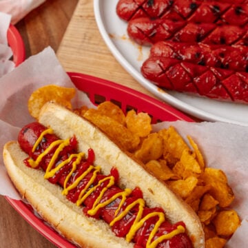 BBQ hotdogs 7 with mustard on red tray, crispy waffle fries, and sliced strawberries on white plate in background