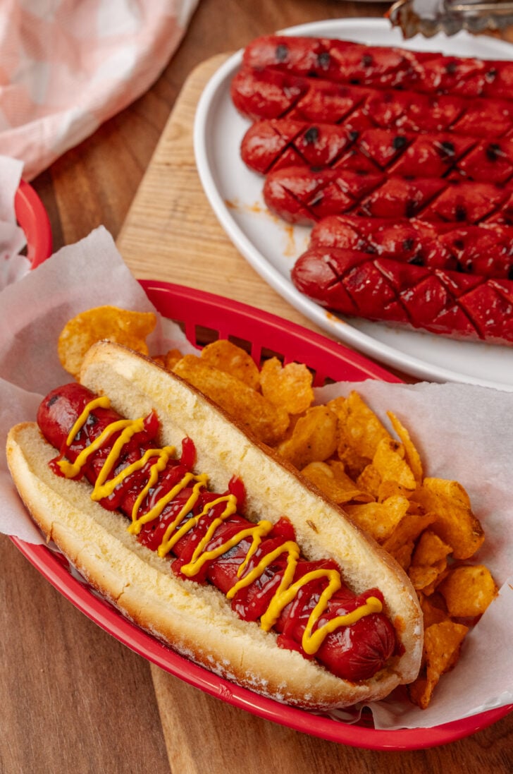 Hot dog in a bun with mustard and ketchup served with chips in a red basket.