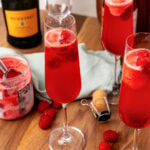 Raspberry mimosas 7 in champagne flutes with fresh raspberries, champagne bottle, and jam jar on wooden board