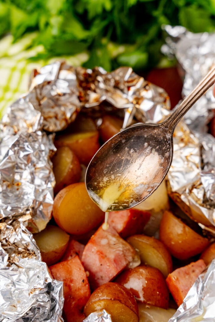 Silver spoon drizzling butter over sausage and potatoes in foil packs.