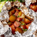 Sausage foil packs 7 with red potatoes, sliced sausage, and fresh parsley garnish on wooden board with fresh herbs