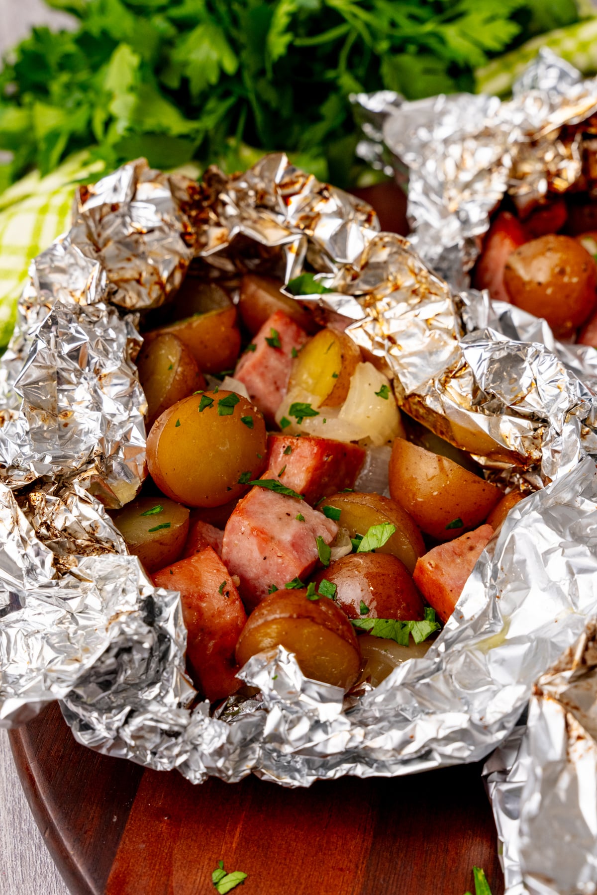 Sausage foil packs 7 with red potatoes, sliced sausage, and fresh parsley garnish on wooden board with fresh herbs