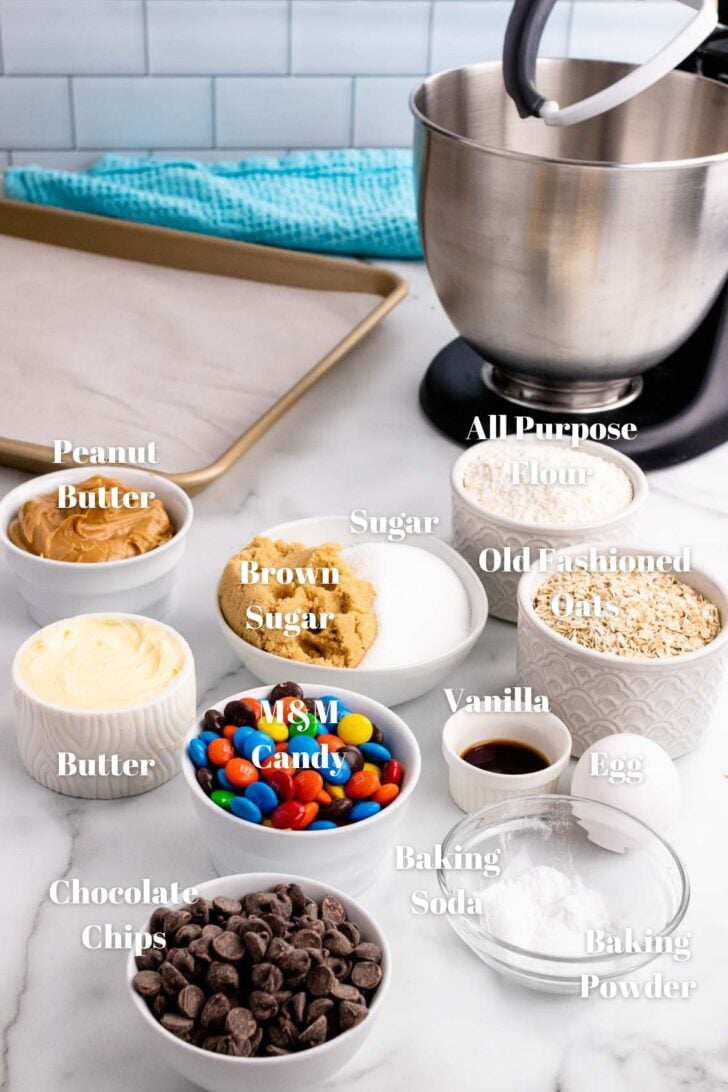 Ingredients for Monster Cookies in glass bowls along with a stand mixer and baking pan.