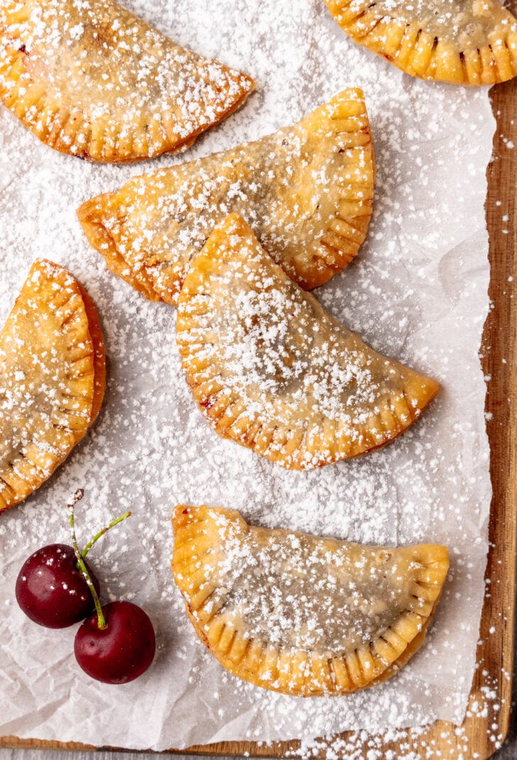 Fried hand pies made with cherries dusted with powdered sugar on parchment paper.
