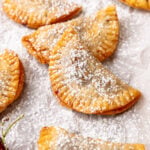 Golden baked cherry pies dusted with powdered sugar on parchment paper with fresh cherries beside them