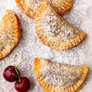 Fried Cherry Pies dusted with powdered sugar on parchment paper with fresh cherries beside them