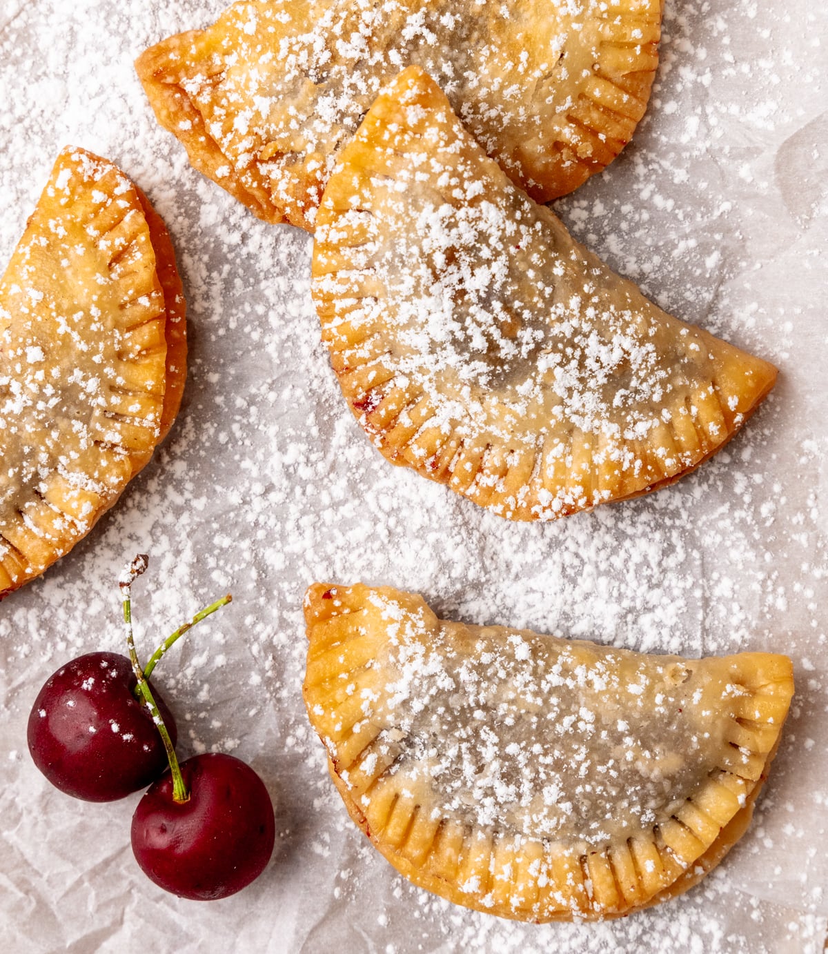 Fried Cherry Pies dusted with powdered sugar on parchment paper with fresh cherries beside them
