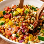 Chickpea Salad with cucumbers, tomatoes, bell peppers, red onion, and feta cheese in white bowl with wooden spoon
