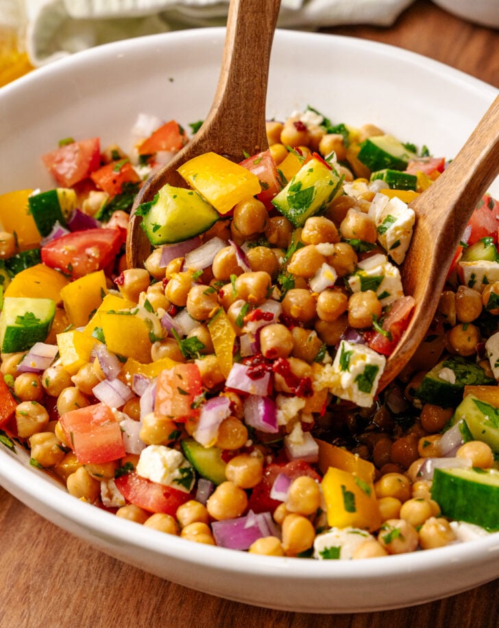 Chickpea Salad with cucumbers, tomatoes, bell peppers, red onion, and feta cheese in white bowl with wooden spoon