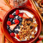 Overhead view of yogurt bowl topped with fresh raspberries, blueberries, strawberries, and homemade granola in a red ceramic dish