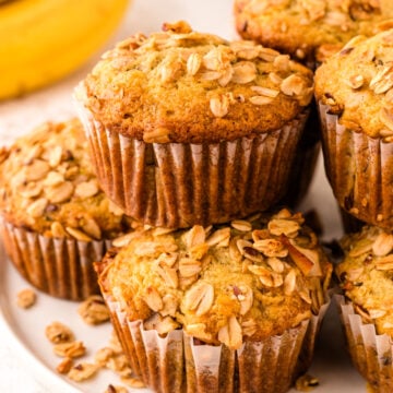 Banana granola muffins topped with oats in brown paper liners arranged on a white plate with fresh bananas blurred in background