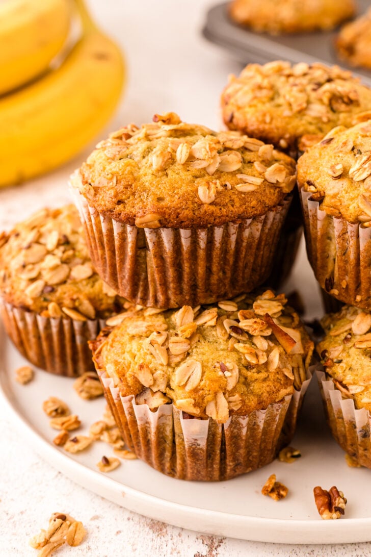 Banana granola muffins topped with oats in brown paper liners arranged on a white plate with fresh bananas blurred in background