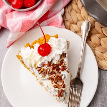Slice of Million Dollar Pie on white plate with whipped cream, pecans, maraschino cherry, fork, and pink checkered napkin nearby