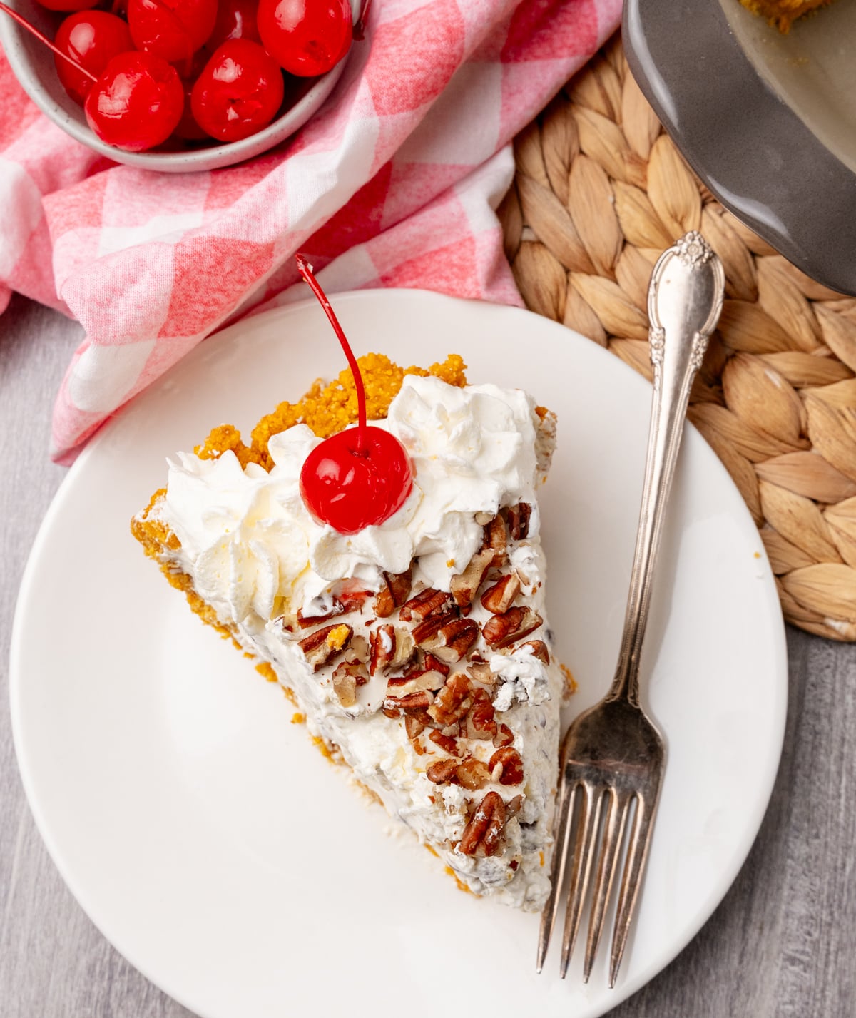 Slice of Million Dollar Pie on white plate with whipped cream, pecans, maraschino cherry, fork, and pink checkered napkin nearby