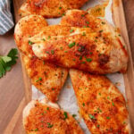 Six seasoned oven chicken breasts on parchment paper with paprika and green onion garnish on wooden cutting board