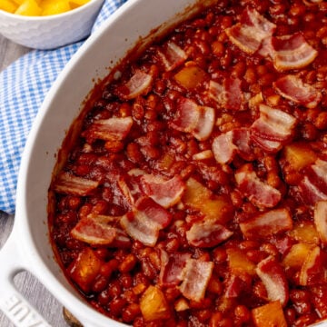 Pineapple baked beans with bacon in white casserole dish, yellow pineapple chunks in bowl beside it on blue gingham cloth
