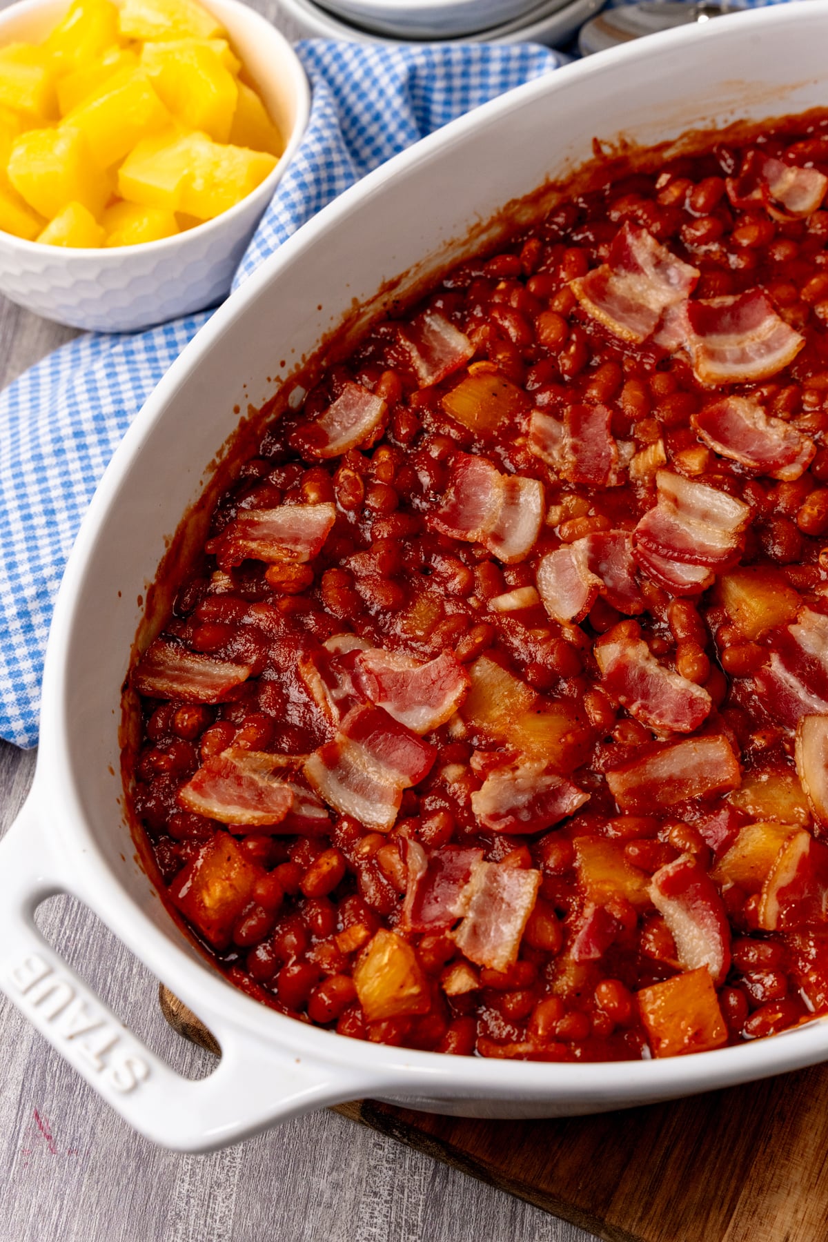 Pineapple baked beans with bacon in white casserole dish, yellow pineapple chunks in bowl beside it on blue gingham cloth