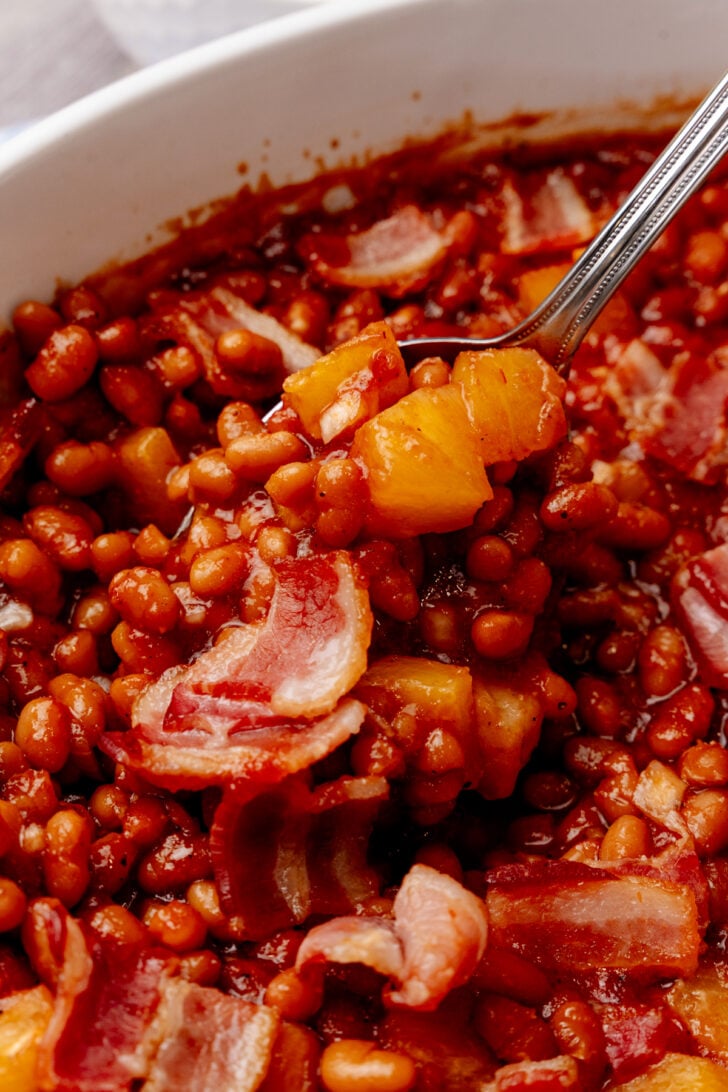 Spoon lifting brown sugar pineapple baked beans with bacon and diced pineapple from white bowl