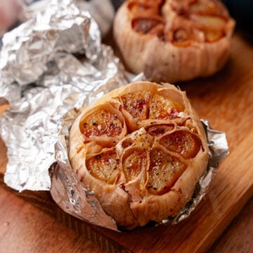 Roasted garlic bulbs wrapped in foil on wooden board, showing caramelized cloves with golden-brown tops and seasoning