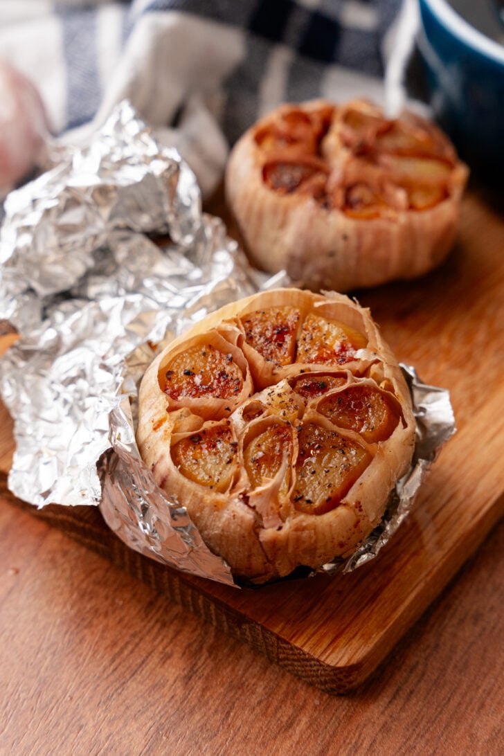 Roasted garlic bulbs wrapped in foil on wooden board, showing caramelized cloves with golden-brown tops and seasoning