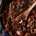 Sauteed mushrooms 9 cooking in a blue Staub cast iron skillet with a wooden spoon, overhead view on gray linen