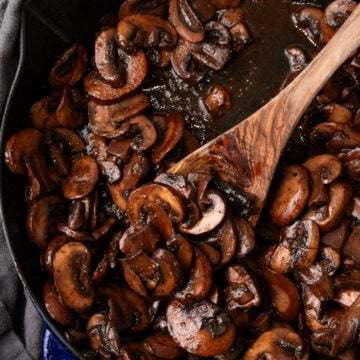 Sauteed mushrooms 9 cooking in a blue Staub cast iron skillet with a wooden spoon, overhead view on gray linen
