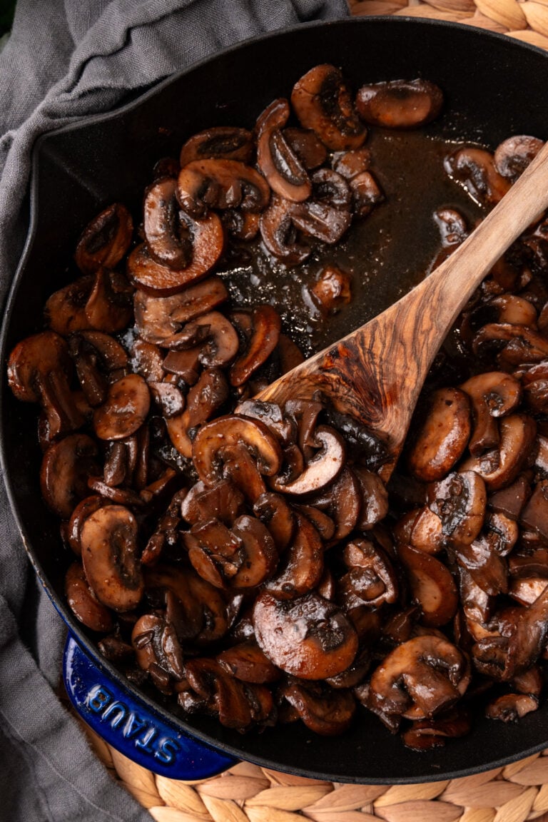 Sauteed mushrooms 9 cooking in a blue Staub cast iron skillet with a wooden spoon, overhead view on gray linen