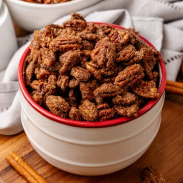 Candied pecans 10 2 in white bowl with red rim on wooden board, cinnamon stick beside, overhead shot with pecan bowls blurred background