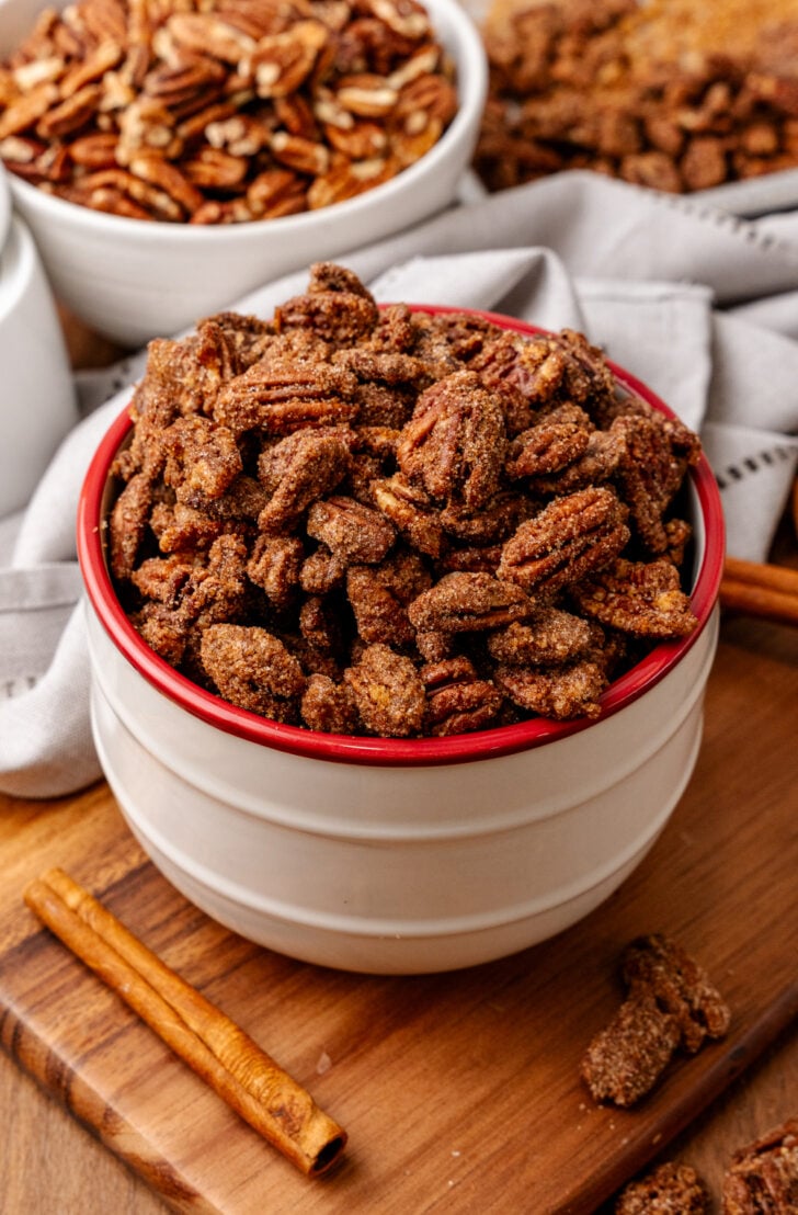 Candied pecans 10 2 in white bowl with red rim on wooden board, cinnamon stick beside, overhead shot with pecan bowls blurred background