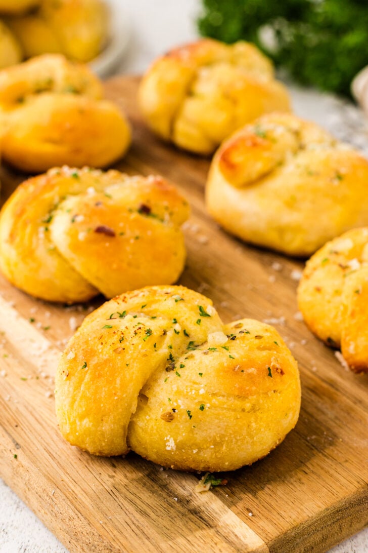 Garlic knots topped with Parmesan cheese and fresh parsley on a wooden cutting board.