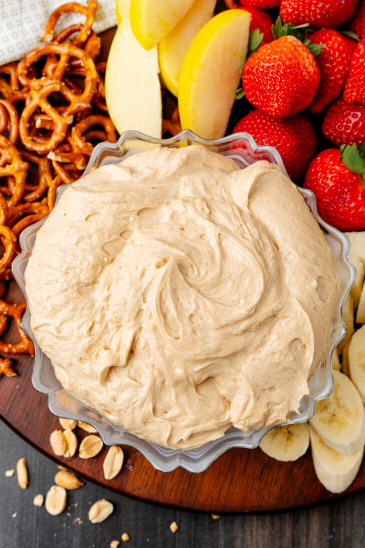Peanut Butter Dip in a scalloped bowl surrounded by pretzels, strawberries, apples, and peanuts on a wooden board