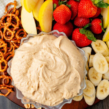 Peanut Butter Dip in a clear bowl surrounded by fresh strawberries, banana slices, pretzel twists, and peanuts on a wooden board