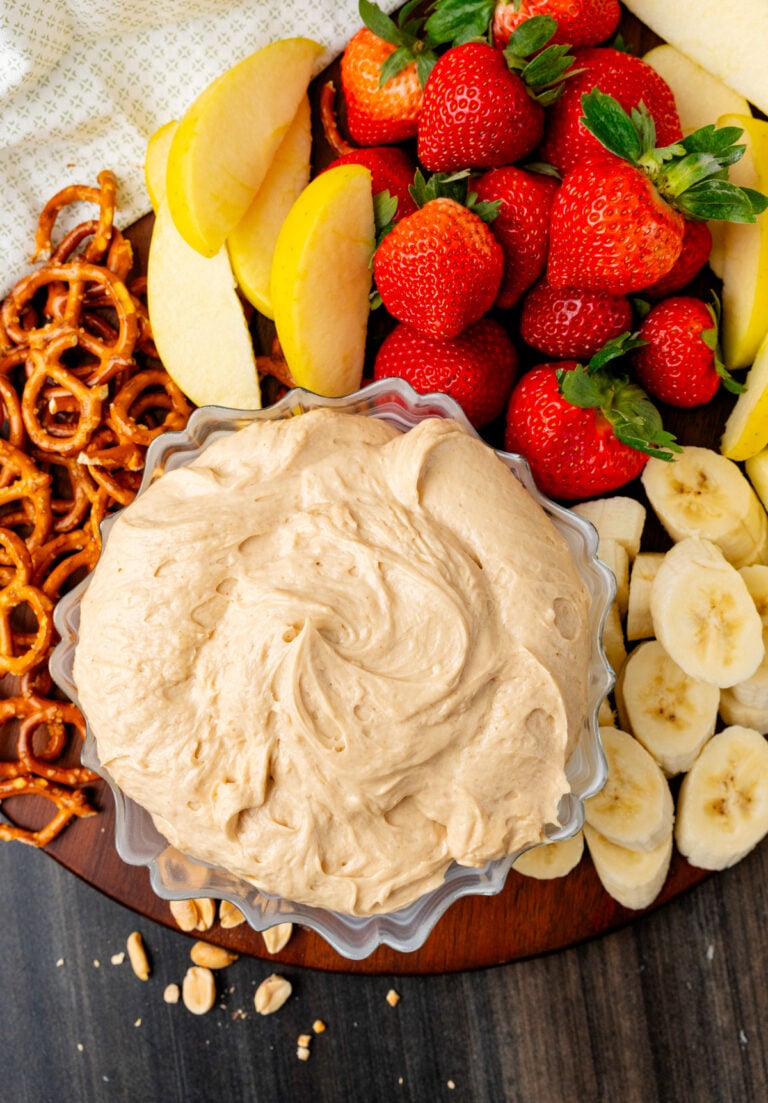 Peanut Butter Dip in a clear bowl surrounded by fresh strawberries, banana slices, pretzel twists, and peanuts on a wooden board