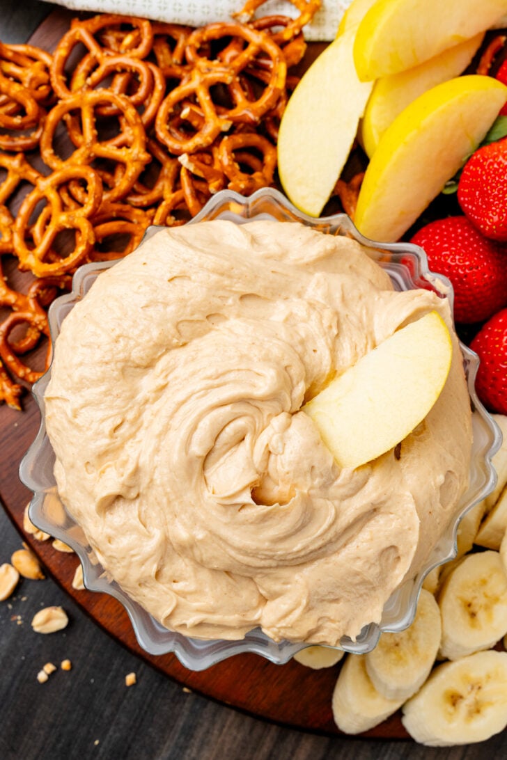 Glass bowl with creamy peanut butter dip served with apple slices, strawberries and pretzels.