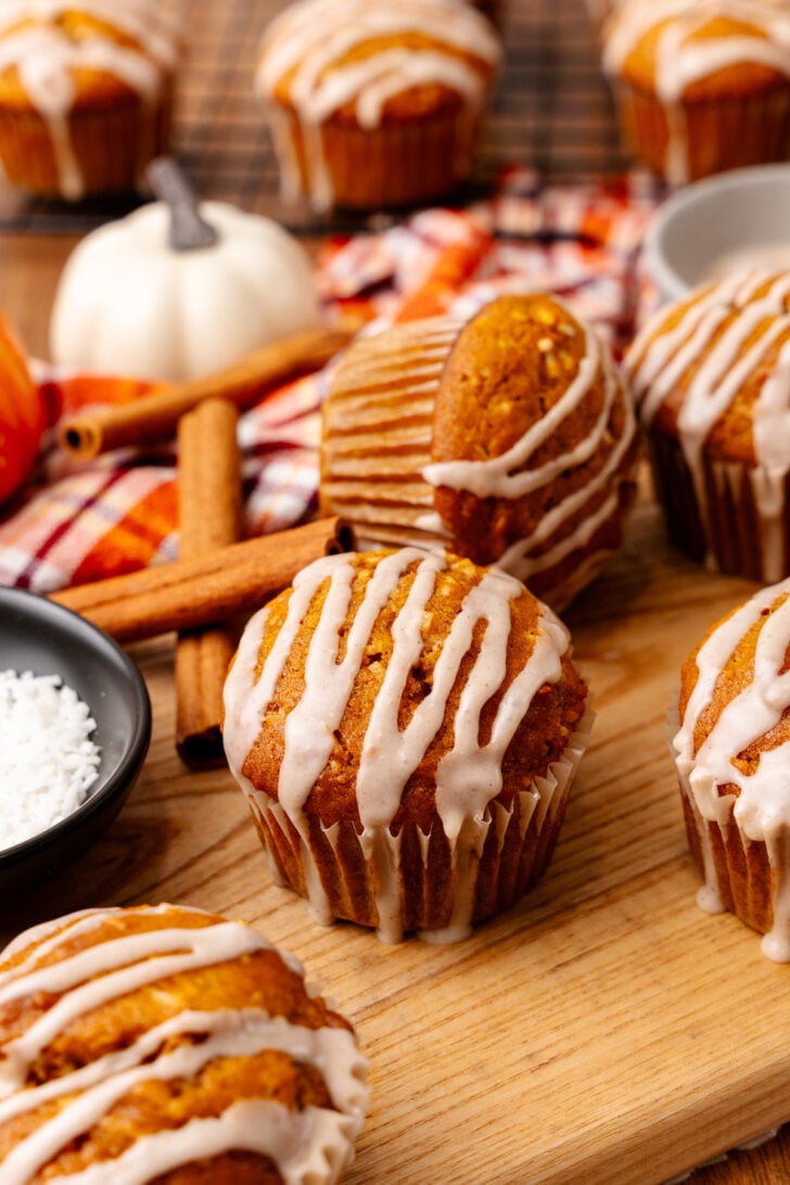 Glazed pumpkin muffins with white icing drizzle in paper liners on wood board with cinnamon sticks and fall decorations