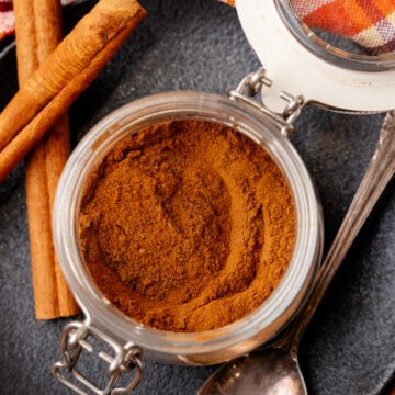Pumpkin spice powder in glass jar with cinnamon sticks and measuring spoons on dark surface with autumn kitchen towel