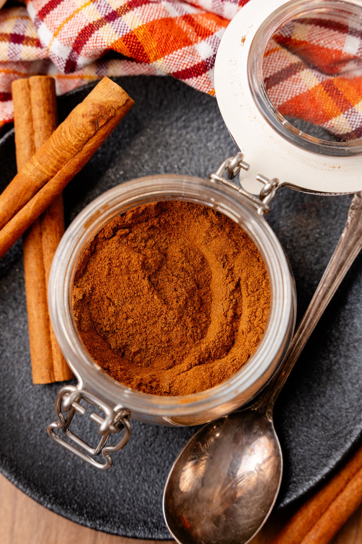 Pumpkin spice powder in glass jar with cinnamon sticks and measuring spoons on dark surface with autumn kitchen towel
