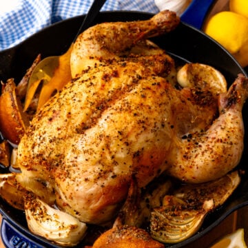 Roasted lemon chicken 4 in cast iron skillet with garlic, onions, and lemon wedges on wooden board