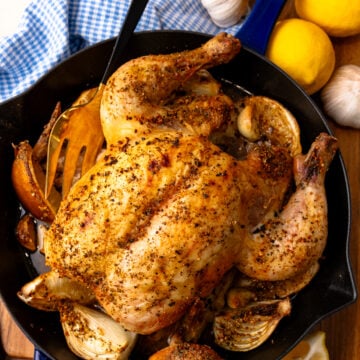 Roasted lemon chicken 5 in cast iron skillet with garlic cloves, lemons, and fresh herbs on wooden table overhead view