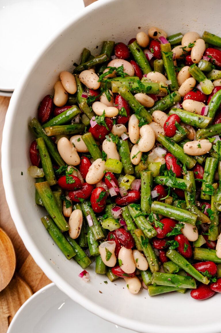 Three bean salad with green beans, white beans, and red peppers garnished with fresh parsley in a white speckled bowl