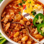 Turkey chili 12 in white bowl topped with sour cream, cheddar cheese, jalapeños, and cilantro with spoon