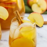 Apple Cider Sangria in glass with ice, apple slices, and cinnamon stick garnish, surrounded by fresh apples