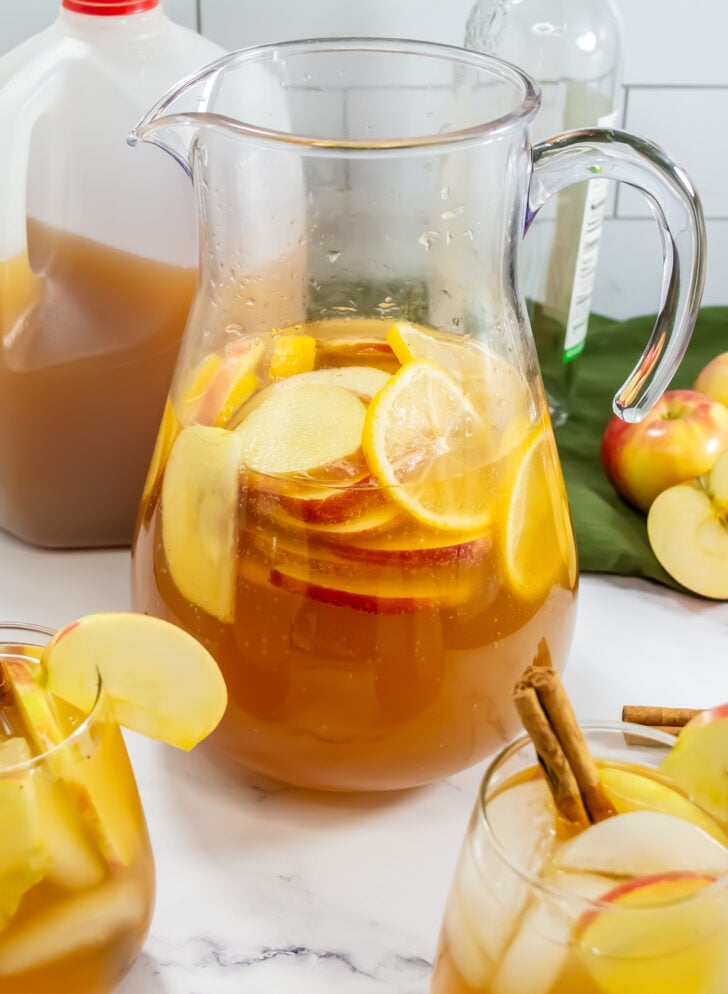 Large glass pitcher filled with apple cider sangria, with lemon and apple slices.