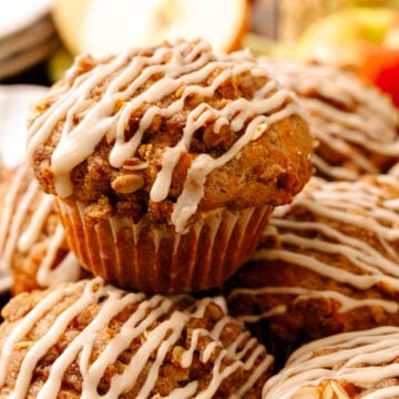 Stack of Honeycrisp Apple Muffins With Maple Glaze drizzled with caramel icing in brown paper liners, surrounded by fresh apples