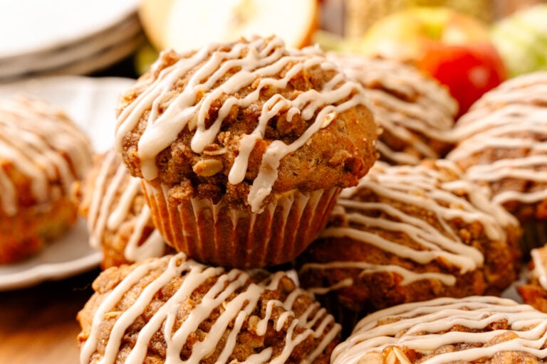 Stack of Honeycrisp Apple Muffins With Maple Glaze drizzled with caramel icing in brown paper liners, surrounded by fresh apples