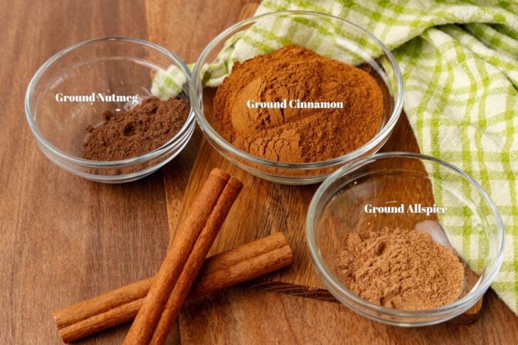 Three glass bowls on a wooden board containing ground spices: nutmeg, cinnamon, and allspice, labeled accordingly. Two cinnamon sticks are placed beside the bowls, with a green checkered cloth in the background.